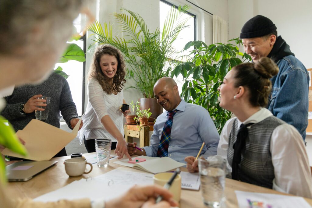 A group of diverse coworkers happily collaborating around a table in a bright, plant-filled office.