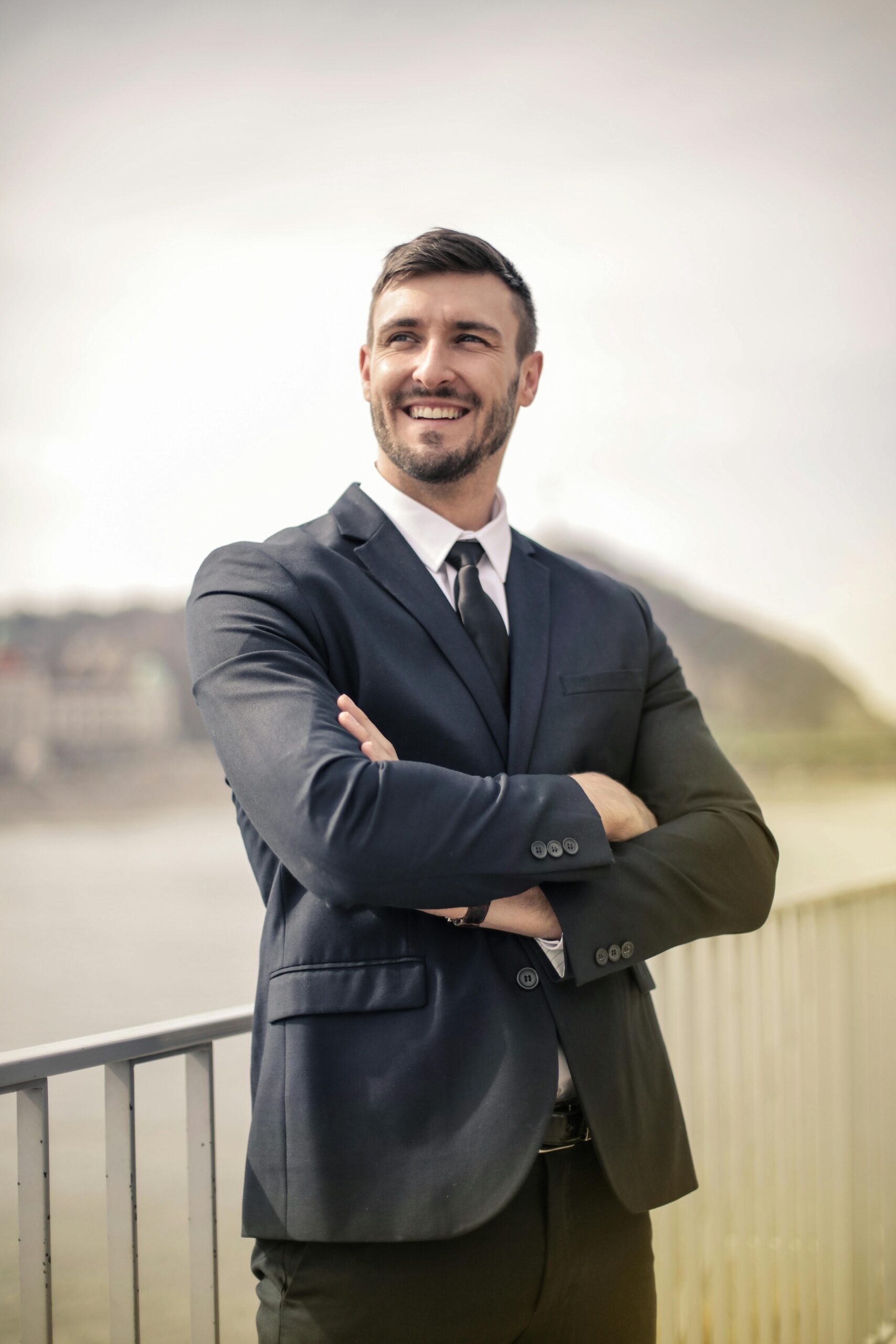 Smiling businessman in a black suit standing outdoors by a railing, showcasing a confident demeanor.